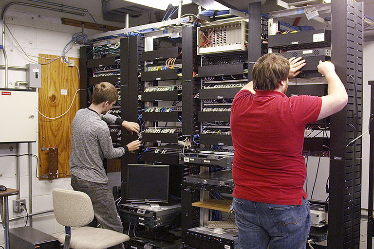 Two individuals in a server room work on a tall rack of networking equipment filled with cables and devices. One wears a gray shirt and stands near a monitor, while the other, in a red shirt, adjusts components higher up. The room features a wooden door, electrical panels, and various cables.